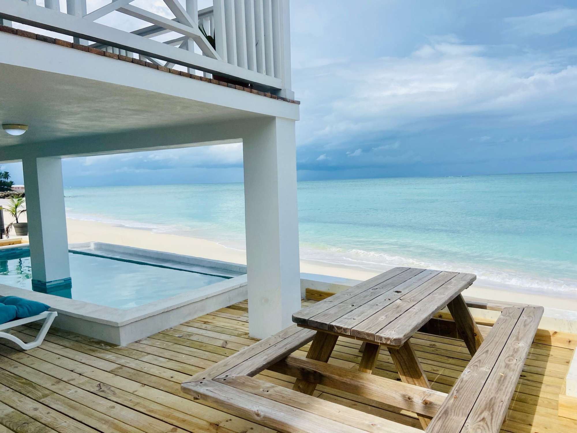 Un pontile in legno con una piscina e un tavolo in legno, con vista su una spiaggia e acque turchesi cristalline, sotto un cielo nuvoloso.