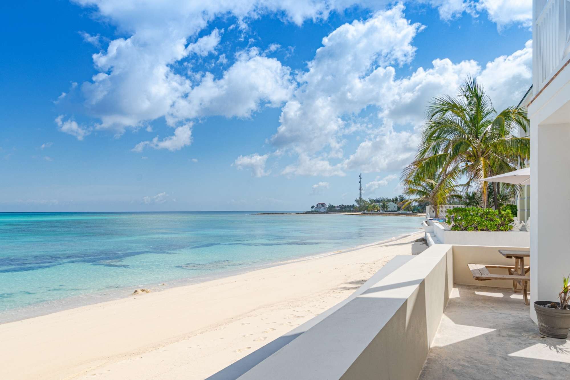 A beach with clear turquoise water, white sand, and a few palm trees. There is a building with a balcony and a potted plant.