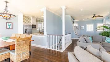 A spacious living area with a dining table and wicker chairs, a staircase leading to an upper level, and a modern kitchen in the background.