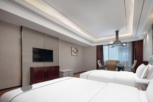 Habitación de hotel con dos camas, un televisor en la pared, una mesa de comedor con sillas y un ventanal con cortinas.