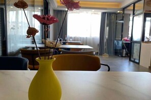 Un vase jaune contenant des fleurs séchées, posé sur une table dans un intérieur moderne, avec de grandes fenêtres donnant sur l’extérieur.