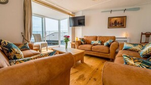 A cozy living room with leather sofas, a wooden coffee table, and a large window with curtains. - The Decker Apartment - East Looe (East Looe)