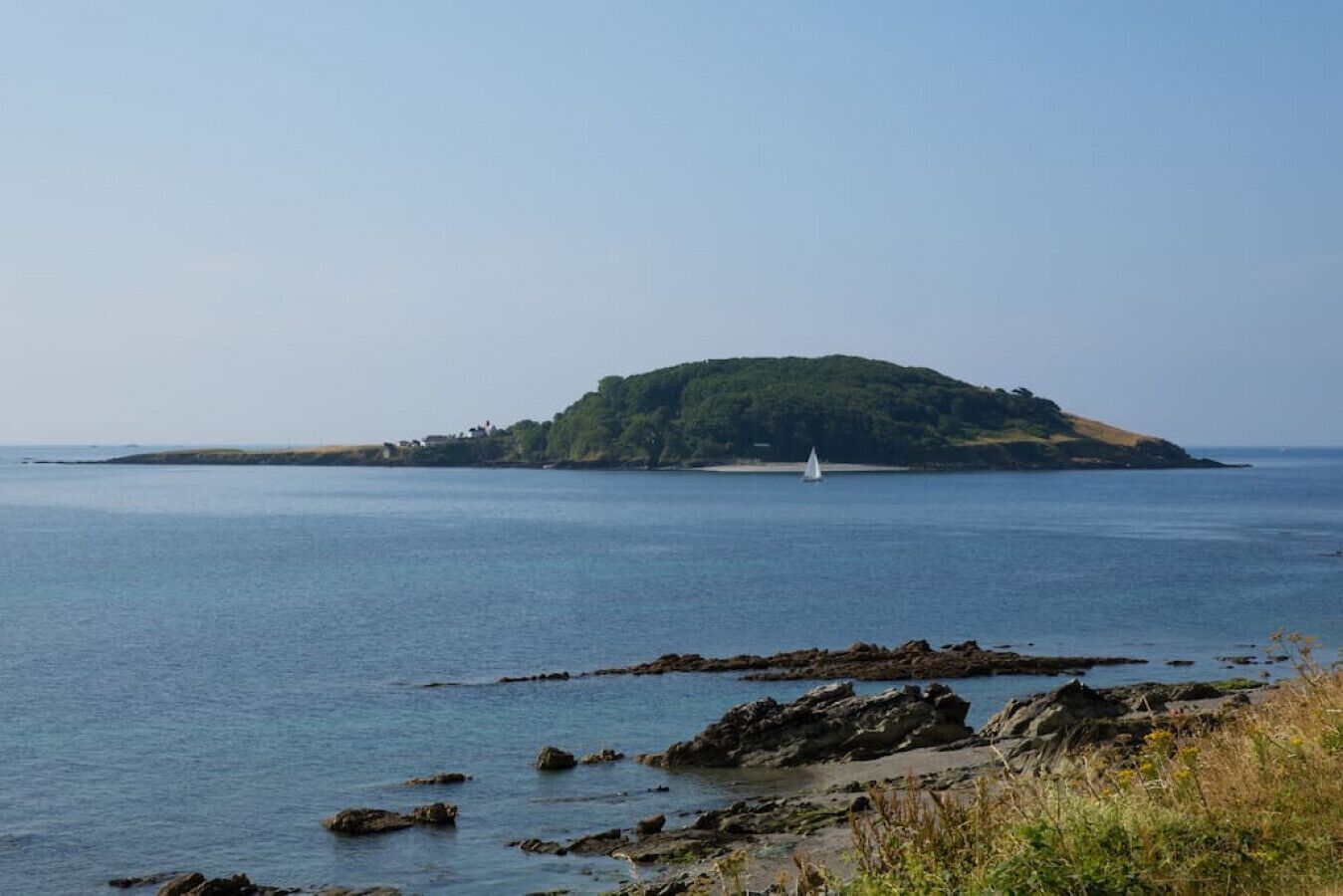 A coastal landscape with a small island, rocky shoreline, and a sailboat in the distance.