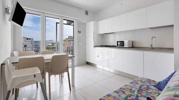 A modern kitchen with white cabinets, a microwave, and a sink. There is a dining area with a table and chairs, and a bed with a colorful quilt. - Apartment "Ca Laurana 42" with Sea View, Air Conditioning and Terrace (Bibione)