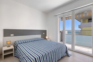 A hotel room with a bed, a nightstand with a lamp, and a sliding glass door leading to a balcony. - Apartment "Condominio Aurora" with Sea View & A/C (Bibione)