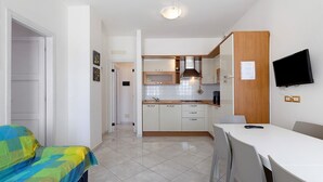 A compact kitchen with white cabinets, a microwave, and a wooden countertop. There is a dining table with chairs and a TV mounted on the wall. - Apartment 'Ca Laurana 52' near the beach with garden and terrace (Bibione)