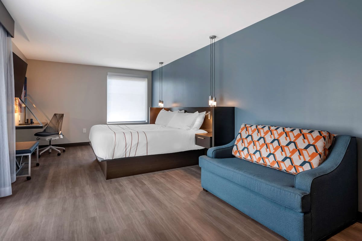 A modern hotel room with a bed, a blue sofa, a desk with a chair, and a window.