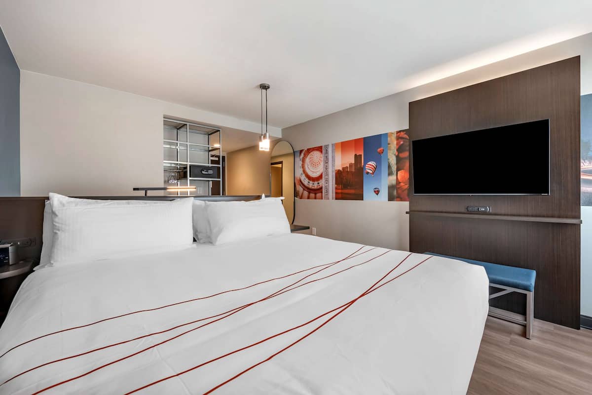 A hotel room with a large bed, a TV mounted on the wall, a desk with a chair, and a bench.