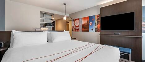 A hotel room with a large bed, a TV mounted on the wall, a desk with a chair, and a bench.