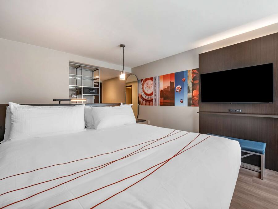 A hotel room with a large bed, a TV mounted on the wall, a desk with a chair, and a bench.