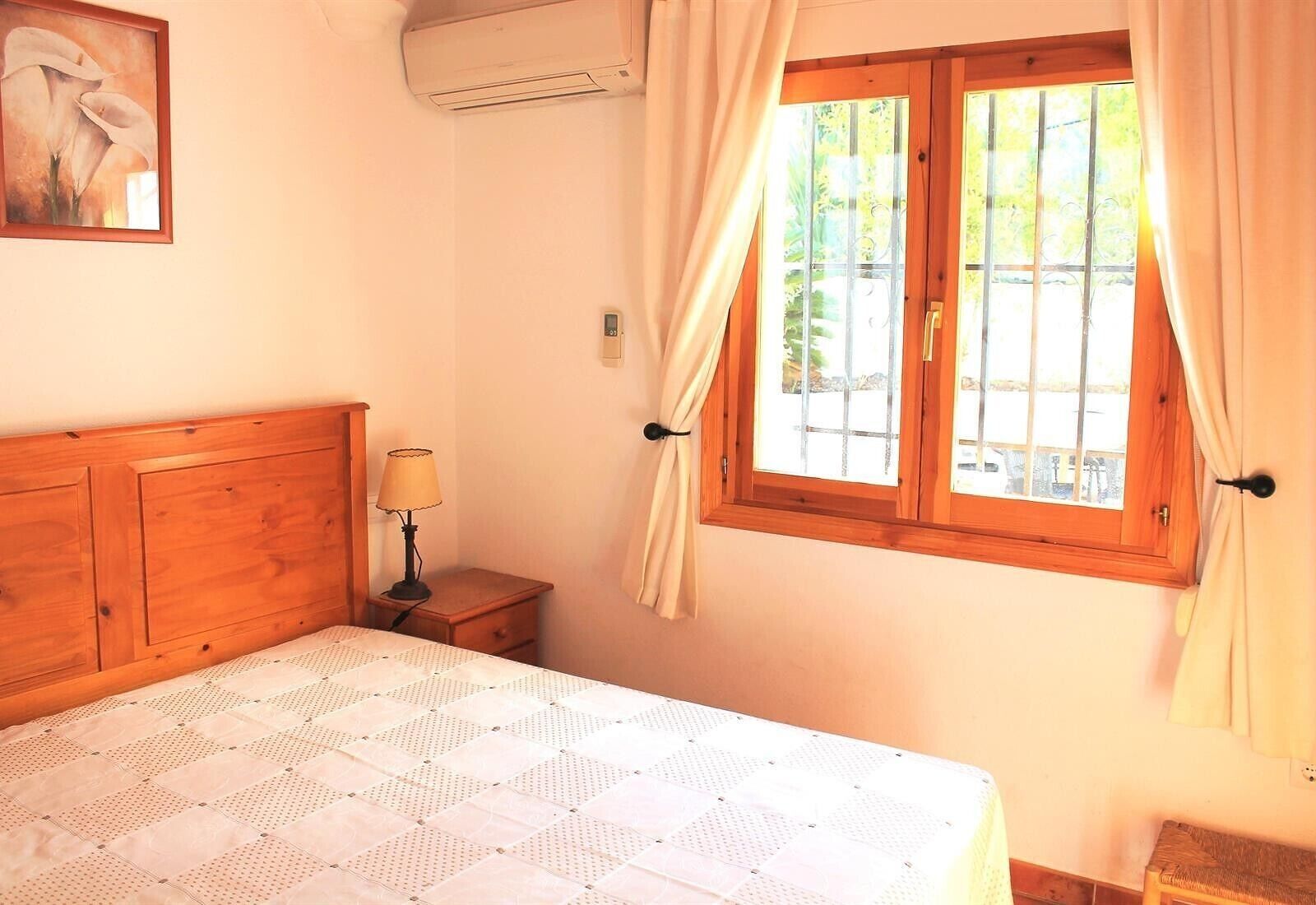 Un dormitorio con cabecera de madera, cama con colcha estampada, lámpara de noche, ventana con cortinas y un equipo de aire acondicionado.