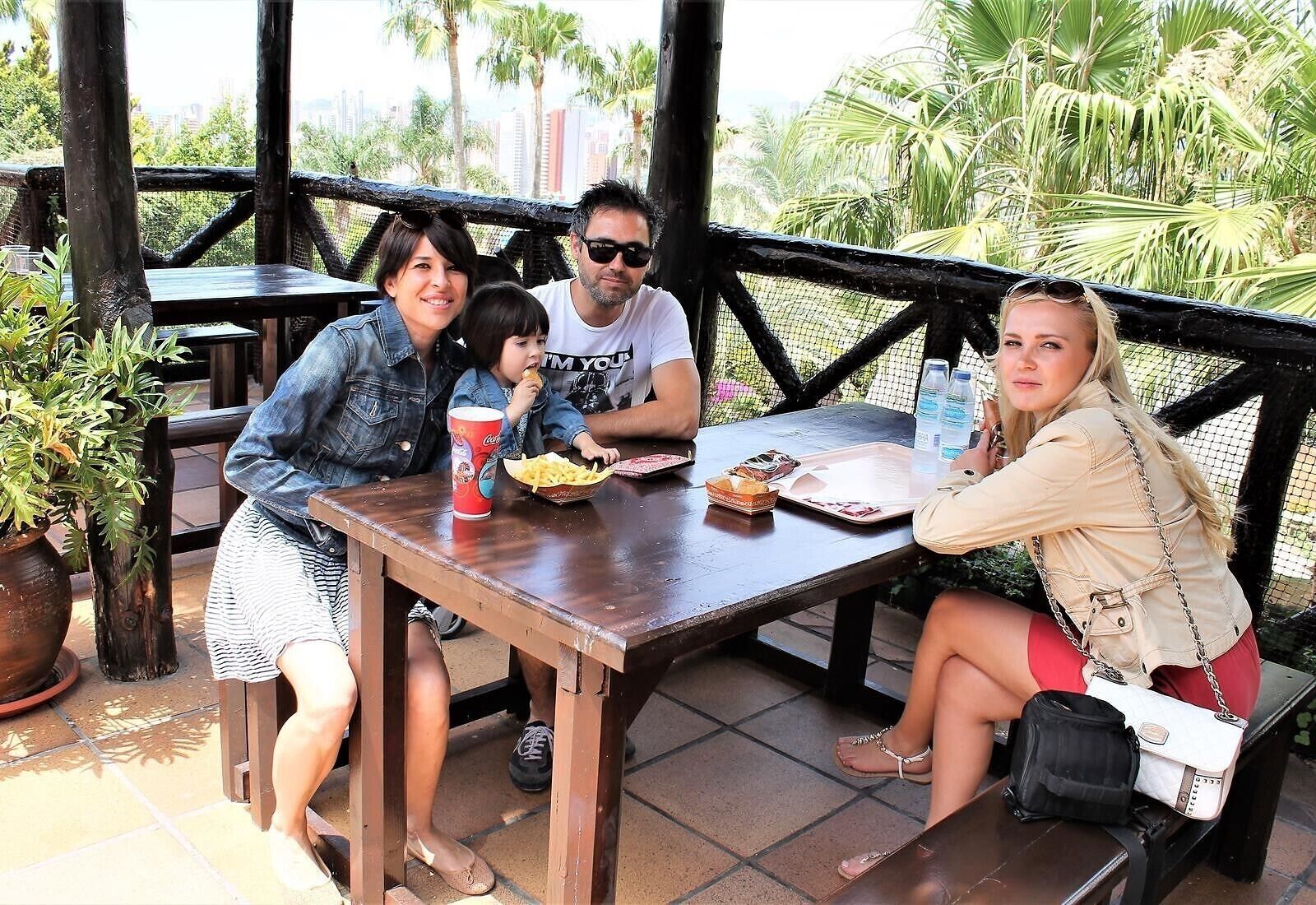 A group of people seated at an outdoor wooden table with a view of palm trees and buildings in the background.