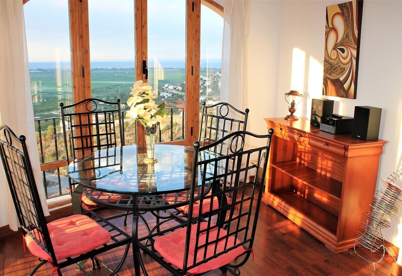 A dining area with a round glass table, black chairs, a vase with flowers, a wooden cabinet with speakers, and a view of the landscape through large windows.
