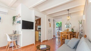 Un salon avec un canapé, un téléviseur, un coin repas avec des chaises, et une cuisine visible au fond.