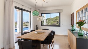 Un comedor moderno con una mesa y sillas de madera, ventanas amplias con vista a una piscina y follaje, y lámparas colgantes.