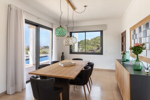 A modern dining area with a wooden table and chairs, large windows with a view of a pool and greenery, and pendant lights.