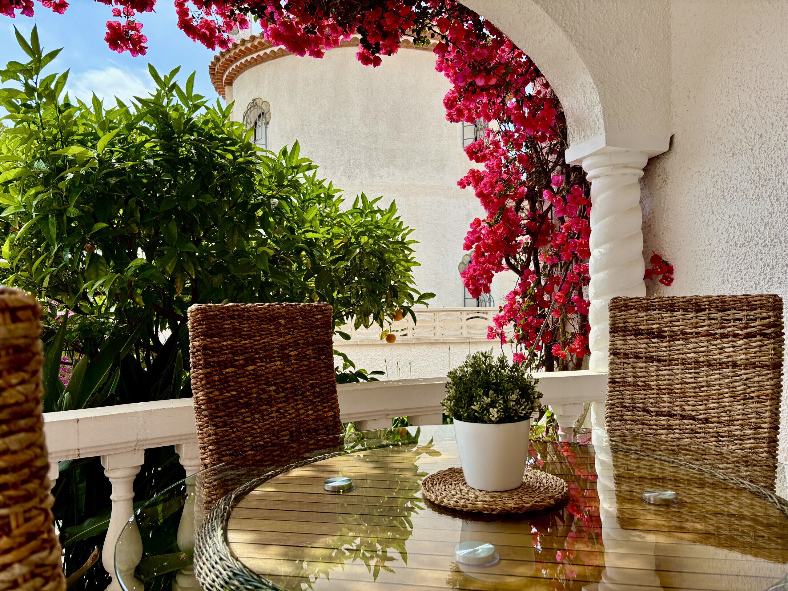 Een terras met rieten stoelen en een glazen tafel, omringd door weelderige begroeiing en kleurrijke bloemen.