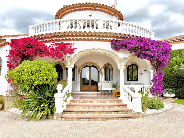 A white building with a balcony, steps leading to a door, and potted plants.
