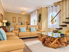 Un salon comprenant un canapé, une table basse en verre avec un socle en bois, un escalier avec une rampe en métal et une vue sur l’extérieur grâce à de grandes fenêtres.