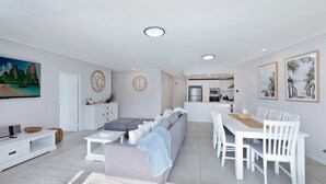 Een moderne woonkamer met een bank, salontafel en televisie. Er is een eethoek met een tafel en stoelen. De keuken is op de achtergrond zichtbaar.