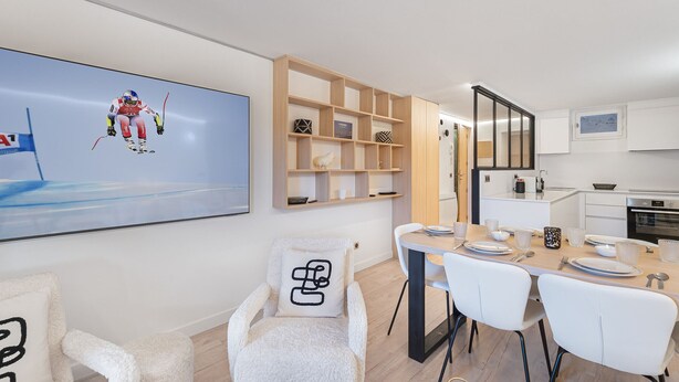 Une cuisine moderne avec des meubles blancs, un coin repas avec des chaises blanches et un grand téléviseur à écran plat fixé au mur.