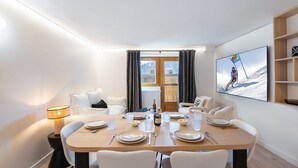 Une salle à manger moderne avec une table en bois dressée pour quatre personnes, des chaises blanches et un téléviseur diffusant une scène de ski.