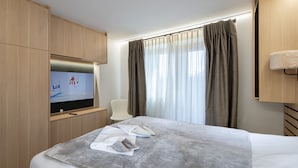 Une chambre d’hôtel avec un lit, un téléviseur fixé à un meuble en bois, une chaise et une fenêtre avec des rideaux.