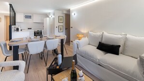 Un salon moderne avec un canapé, une table à manger et des chaises, une cuisine en arrière-plan, et une petite table avec une bouteille et des verres.