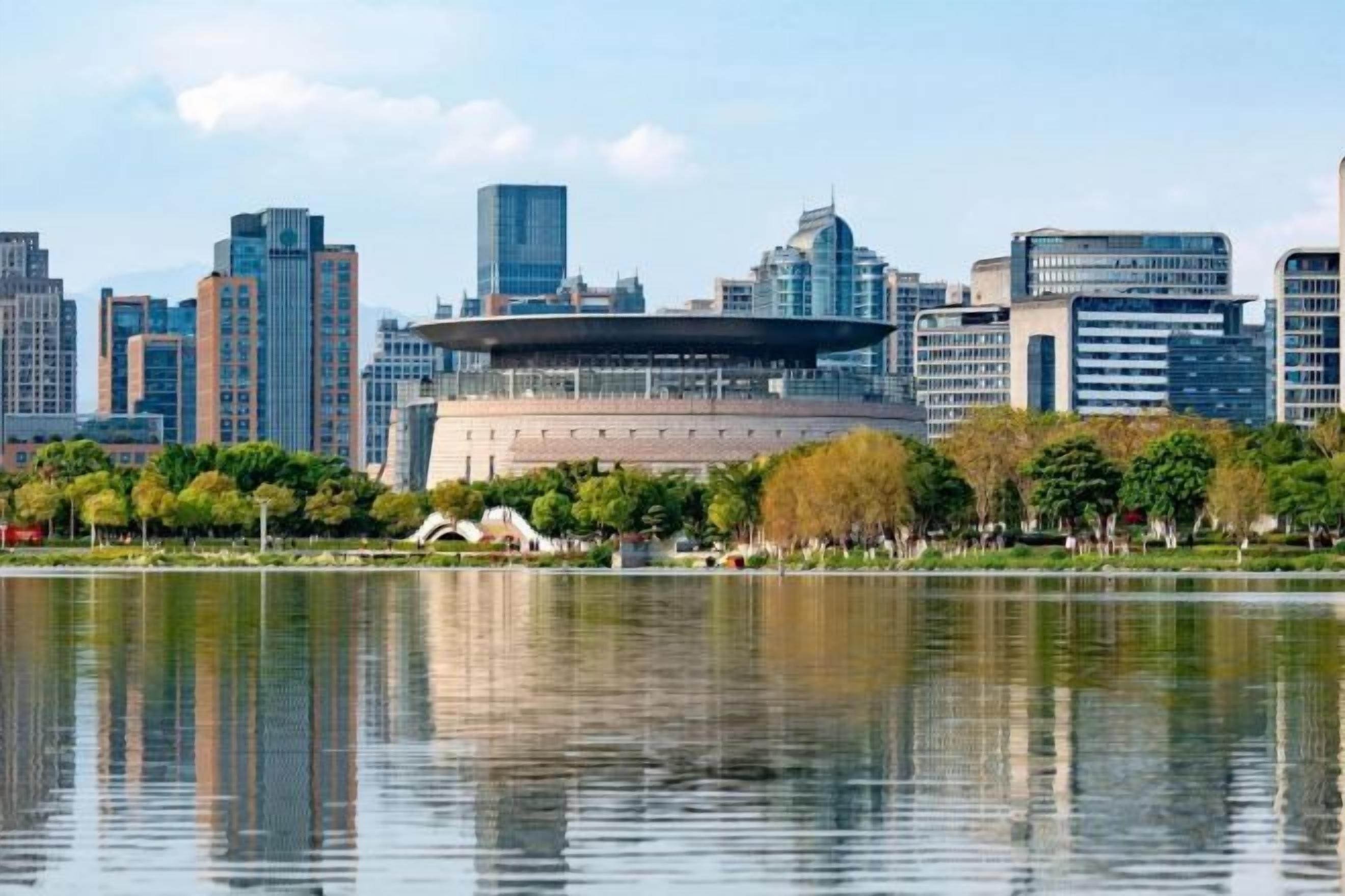 Un paysage urbain avec des bâtiments modernes et une grande structure circulaire au centre, qui se reflète dans une étendue d'eau calme.
