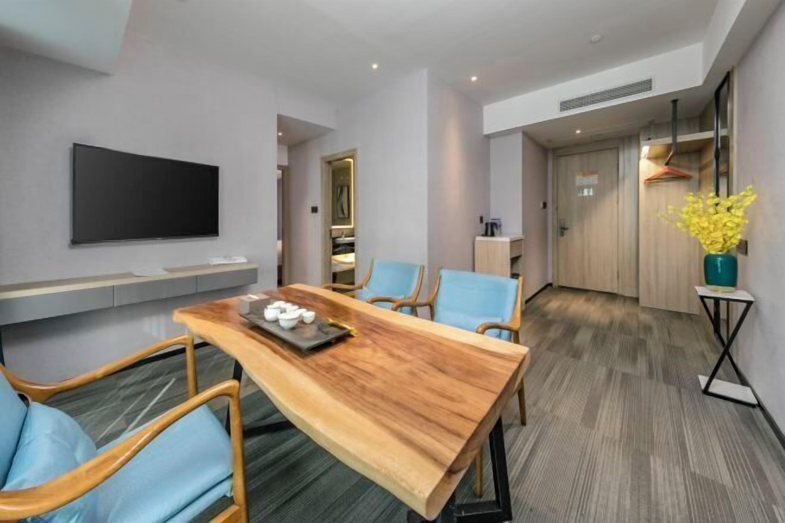 Ein modernes Hotelzimmer mit einem Holzespeisetisch, blauen Stühlen, einem flachbildfernseher an der Wand und einem kleinen Beistelltisch mit einer Vase gelber Blumen.