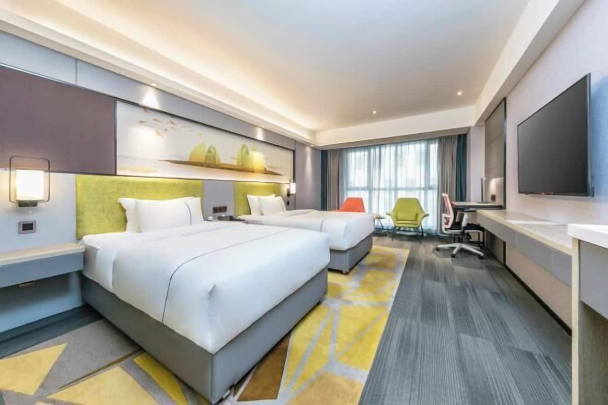 A hotel room with two beds, a desk, a chair, a TV, and a large window with curtains.