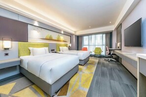 Habitación de hotel con dos camas, un escritorio, una silla, un televisor y un ventanal grande con cortinas.