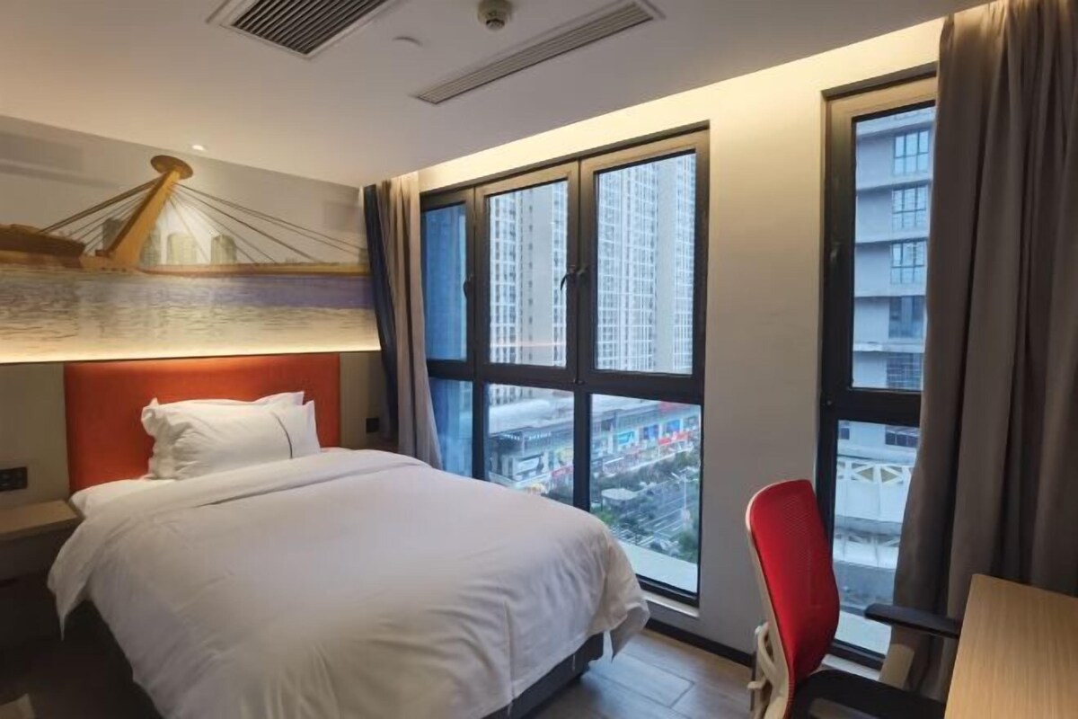 A hotel room with a bed, a red chair, a large window with a city view, and a wall-mounted lamp.