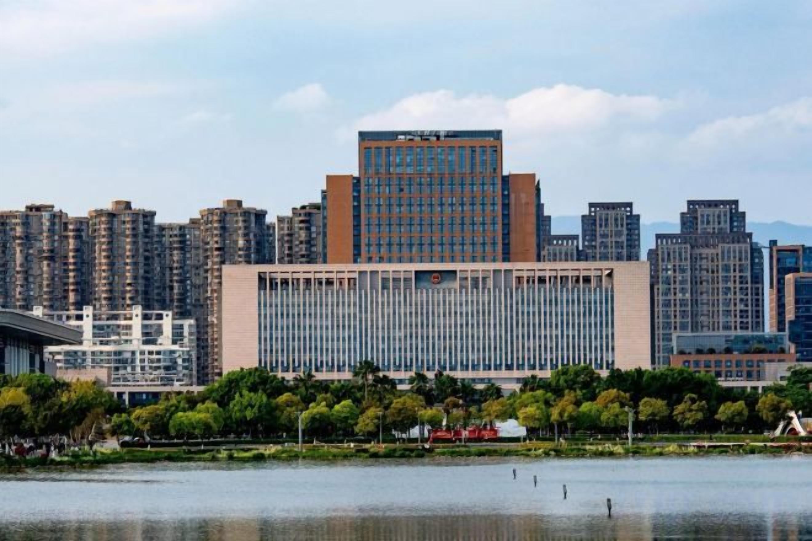 A cityscape with a prominent building in the center, surrounded by high-rise buildings, a body of water, and a park area with trees.