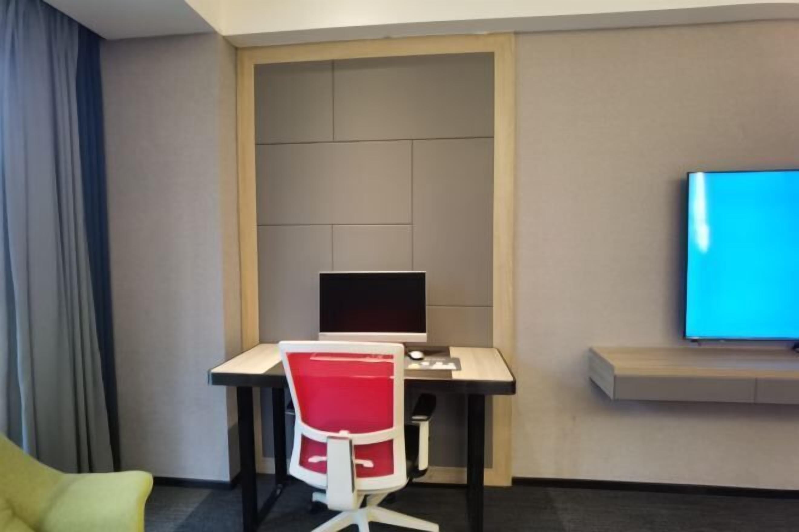 A hotel room with a desk, a red chair, a flat-screen TV, and a window with curtains.