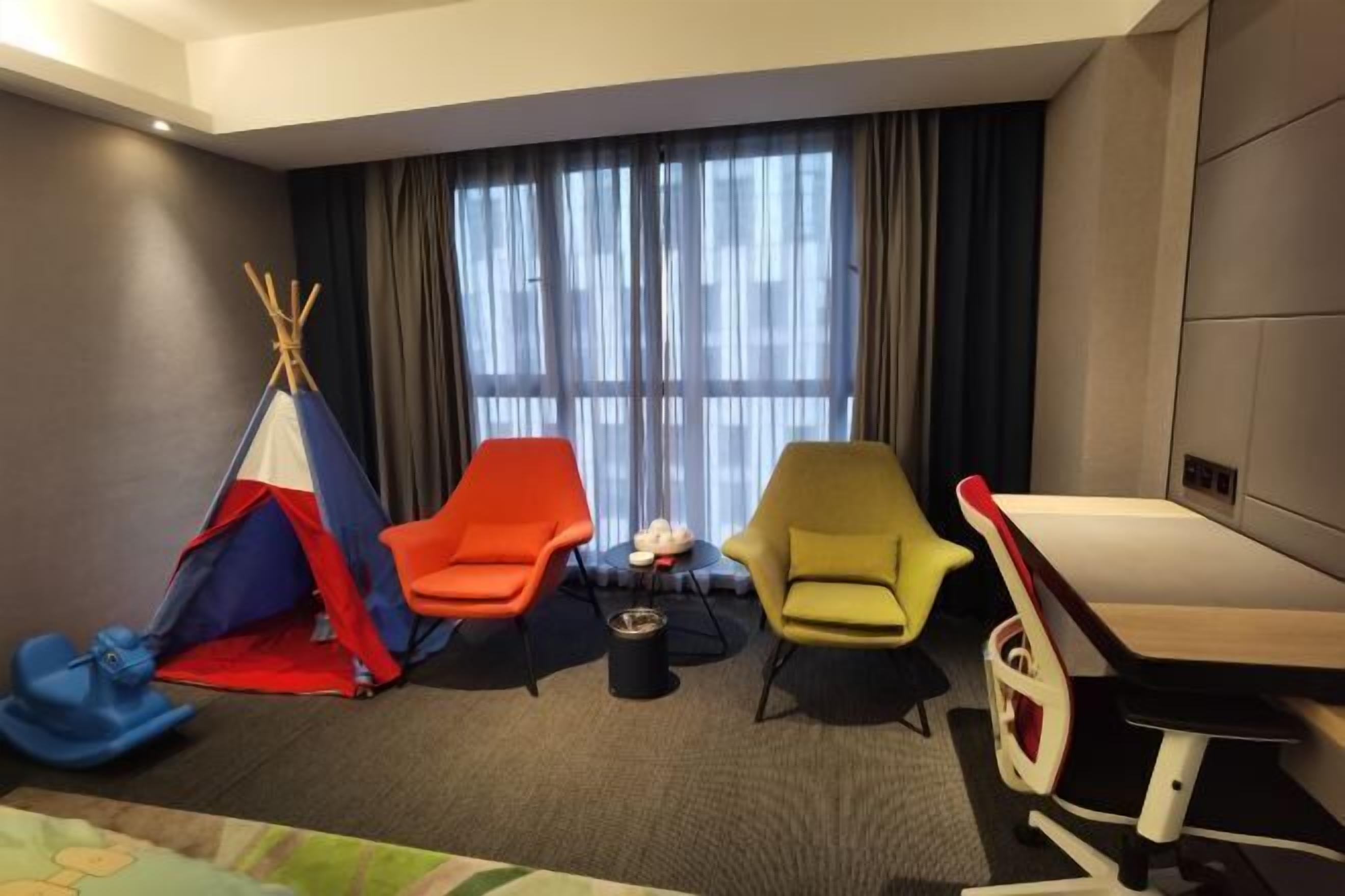 Een hotelkamer met een groot raam, een bed, een bureau en kleurrijke stoelen.