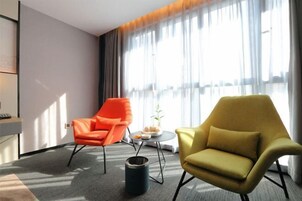Una habitación de hotel moderna con dos sillones, una mesita redonda y un ventanal con cortinas translúcidas.
