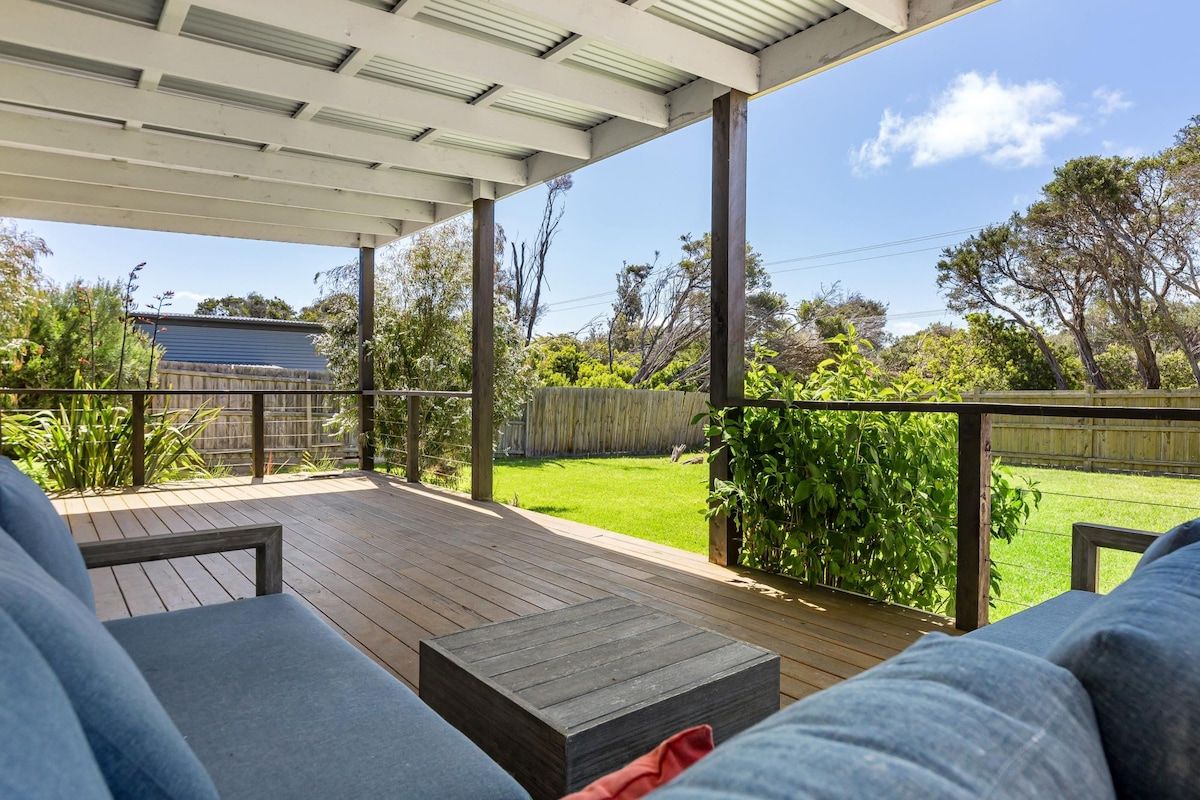A covered outdoor area with a wooden deck, seating, and a view of a backyard with a lawn and trees.