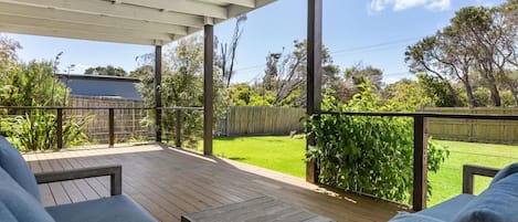 A covered outdoor area with a wooden deck, seating, and a view of a backyard with a lawn and trees.