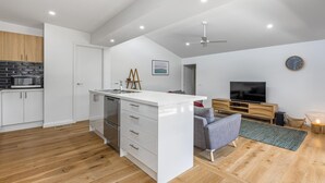 A modern kitchen with white cabinetry, a built-in microwave, and a dishwasher. There is a dining area with a grey sofa and a wooden TV stand.