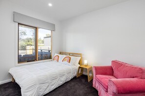 A bedroom with a bed, a nightstand, a red sofa, and a window with a view of the outside.