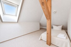 Une chambre avec un plafond en pente, une lucarne, un lit avec une literie blanche et une structure en poutres apparentes.