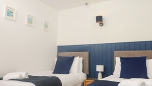 Habitación de hotel con dos camas, ropa de cama blanca, cabeceros azul oscuro y una mesita de noche pequeña con una lámpara.