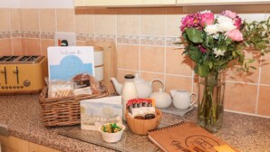 A kitchen counter with a vintage toaster, a wicker basket, a card, a vase with flowers, a wooden bowl with jars, and a notebook. - Primrose Cottage (Paignton)