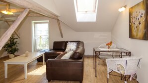 Un espacio tipo loft con techo inclinado, un sofá de cuero marrón, una mesa de centro de vidrio, una silla de mimbre blanco y un cuadro en la pared.