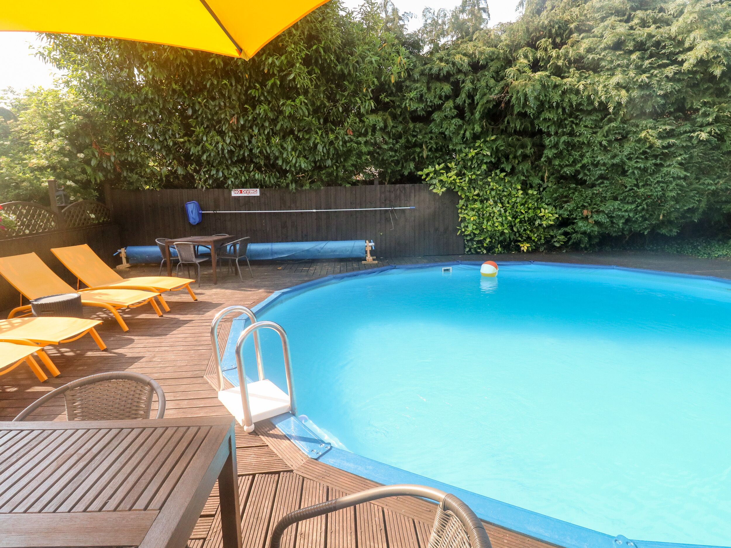 Uma piscina com espreguiçadeiras e uma mesa, cercada por um deck de madeira e um sebe.