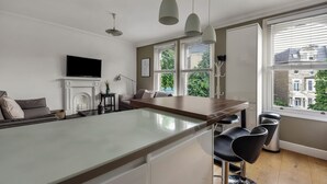 A modern kitchen with a central island, bar stools, a fireplace, and large windows offering views of the neighborhood. - Immaculate 2 Bed Flat in West Kensington (London)
