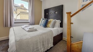 A neatly made bed with a white and gray herringbone throw blanket, a dark headboard with circular details, and a view of a building through a window with curtains. - Immaculate 2 Bed Flat in West Kensington (London)