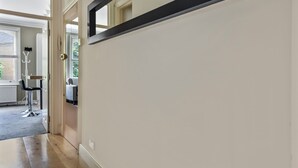 A hallway with a door leading to a room. There is a coat rack with a few items on it, and a chair is visible in the background. - Immaculate 2 Bed Flat in West Kensington (London)