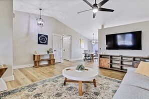 A living room with a round coffee table, a flat-screen TV, a ceiling fan, and a dining area with a wooden table and chairs. - Calming 3 bedroom home with designer details in Na (Nampa)